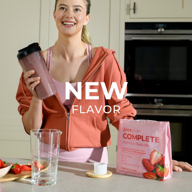 Woman holding a juice plus+ Complete shake mix package and a shake in a kitchen.