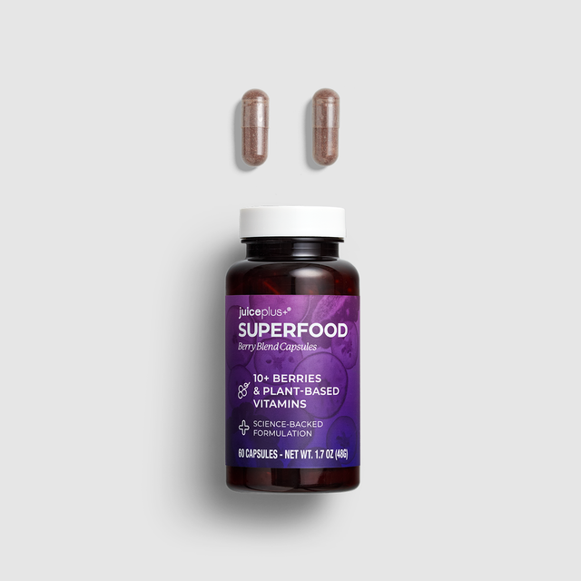 A bottle of juiceroom+ Superfood Berry Blend Capsules with two brown capsules floating above it on a light gray background.