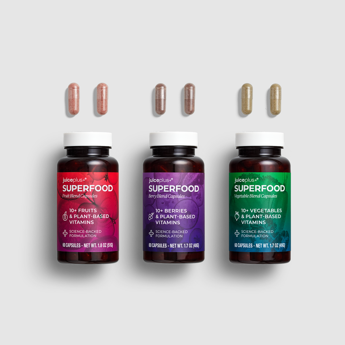Three JuicePlus+ Superfood supplement bottles in red, purple, and green, shown with matching capsules above each bottle on a light gray background.