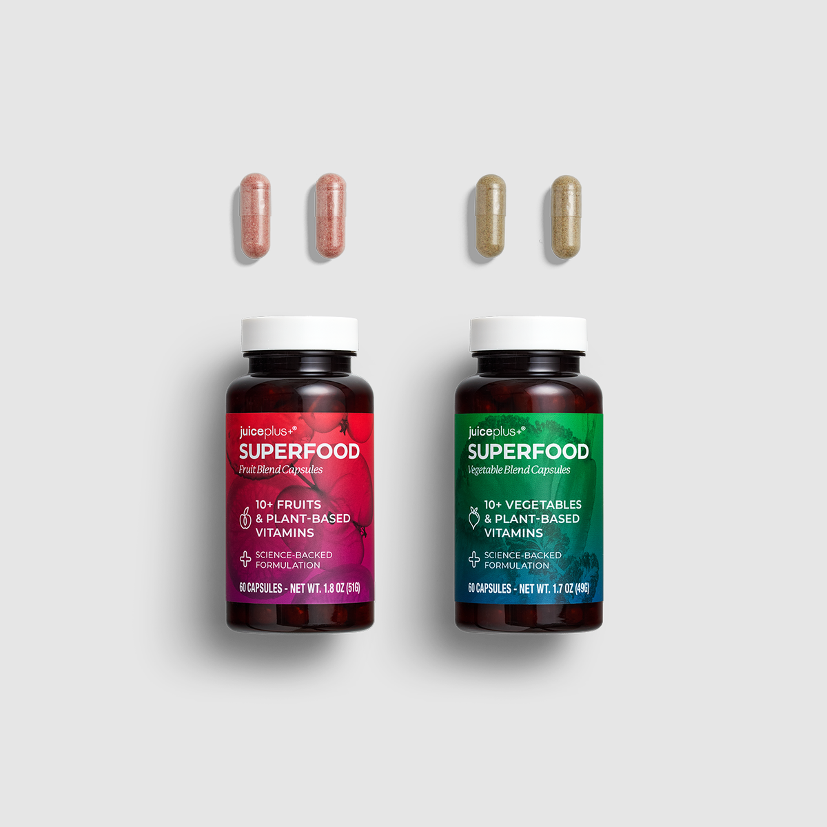 Two JuicePlus+ Superfood supplement bottles on a light gray background, each with three capsules floating above it. The left bottle has a pink label and the right bottle has a green label.